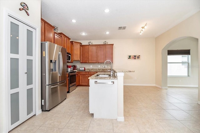 kitchen with arched walkways, brown cabinetry, an island with sink, appliances with stainless steel finishes, and a sink