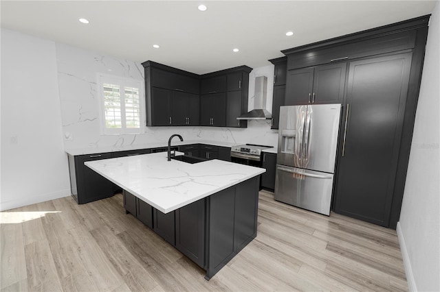 kitchen featuring appliances with stainless steel finishes, a kitchen island with sink, light stone counters, wall chimney range hood, and light hardwood / wood-style flooring