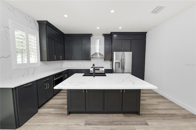 kitchen featuring wall chimney exhaust hood, tasteful backsplash, light stone counters, an island with sink, and stainless steel appliances