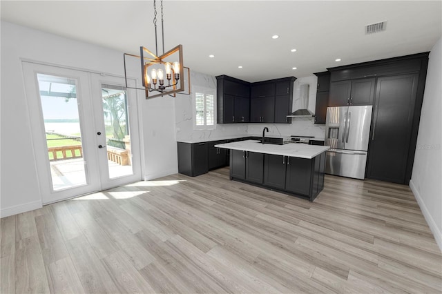 kitchen with wall chimney range hood, stainless steel fridge, a notable chandelier, a center island with sink, and french doors