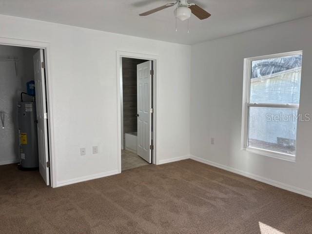 unfurnished bedroom with ceiling fan, a spacious closet, gas water heater, ensuite bathroom, and dark carpet