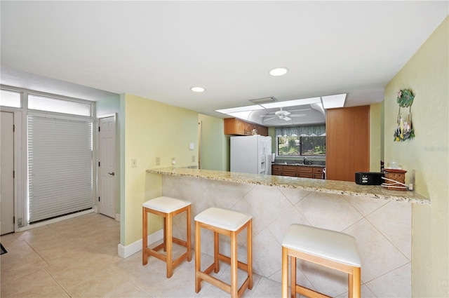 kitchen featuring white refrigerator with ice dispenser, kitchen peninsula, light stone countertops, a kitchen bar, and ceiling fan