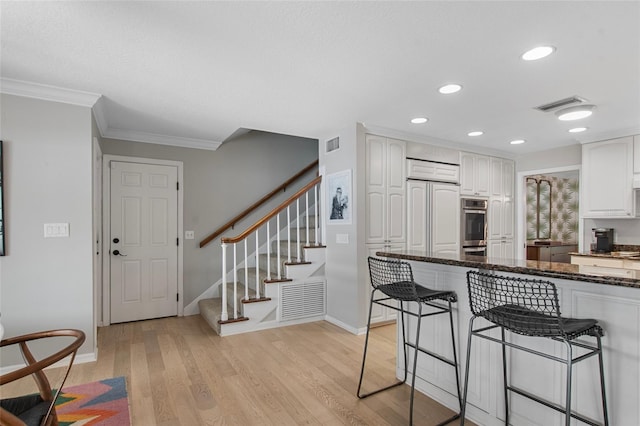 kitchen with white cabinetry, stainless steel double oven, light hardwood / wood-style flooring, dark stone counters, and a breakfast bar