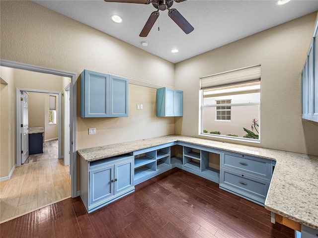 kitchen with dark wood finished floors, built in desk, blue cabinetry, recessed lighting, and light stone countertops