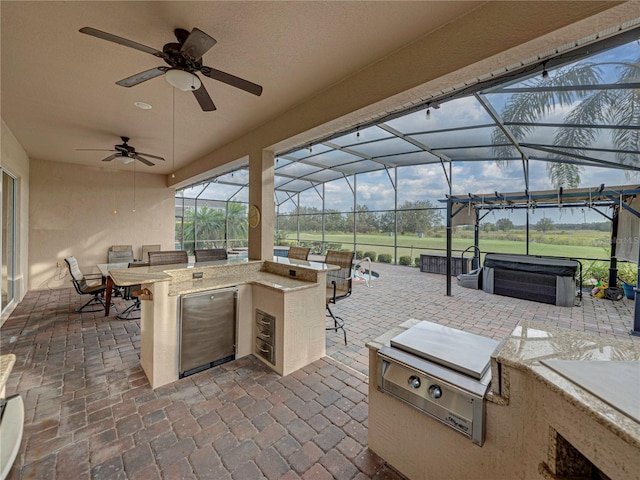 view of patio featuring a hot tub, a ceiling fan, glass enclosure, exterior kitchen, and an outdoor bar