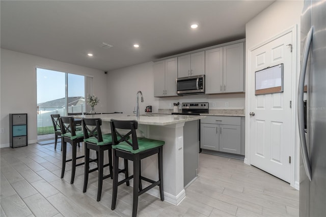 kitchen with gray cabinetry, a kitchen island with sink, light stone countertops, appliances with stainless steel finishes, and a kitchen bar