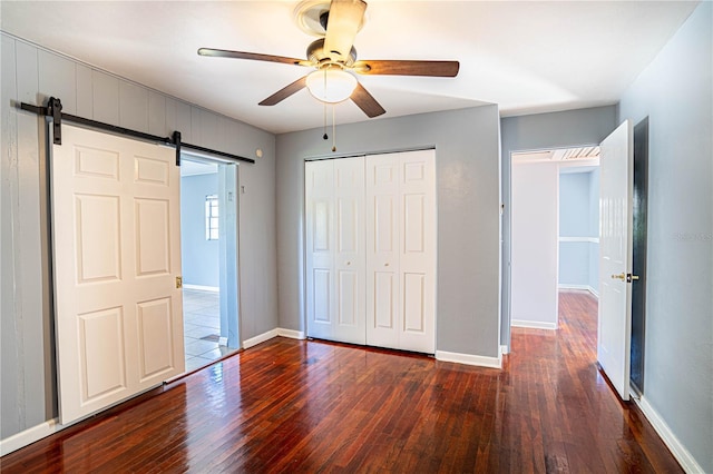 unfurnished bedroom with a barn door, ceiling fan, a closet, and dark wood-type flooring