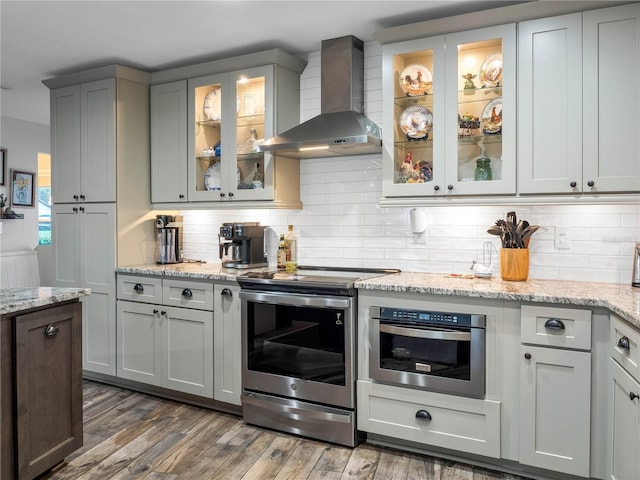 kitchen with wall chimney exhaust hood, light stone counters, dark hardwood / wood-style flooring, backsplash, and appliances with stainless steel finishes