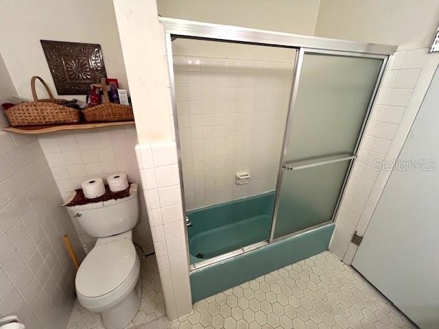 bathroom featuring tile patterned flooring, toilet, shower / bath combination with glass door, and tile walls