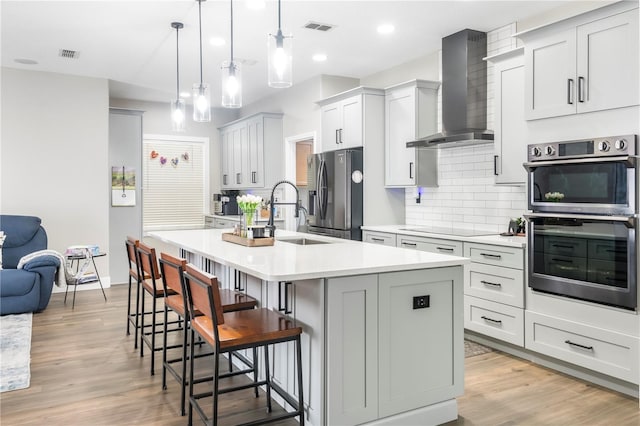 kitchen featuring double oven, a center island with sink, decorative light fixtures, and wall chimney range hood