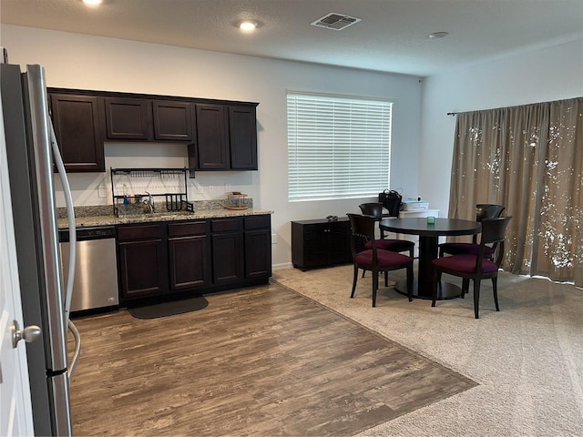 kitchen with wood-type flooring, appliances with stainless steel finishes, light stone countertops, dark brown cabinetry, and sink