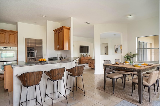 kitchen featuring stainless steel refrigerator with ice dispenser, light tile patterned flooring, a kitchen breakfast bar, kitchen peninsula, and double oven