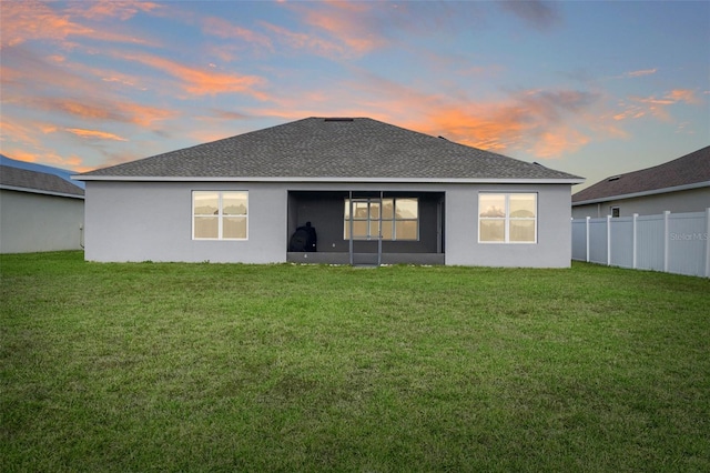 back of property at dusk with stucco siding, roof with shingles, fence, and a yard