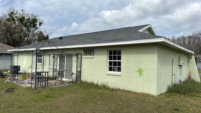 rear view of property with roof with shingles, concrete block siding, a lawn, and a patio