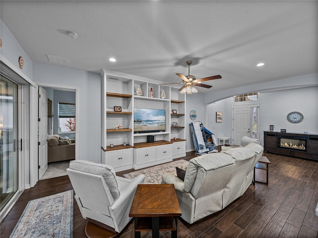 living area with a textured ceiling, dark wood-type flooring, visible vents, a ceiling fan, and a glass covered fireplace