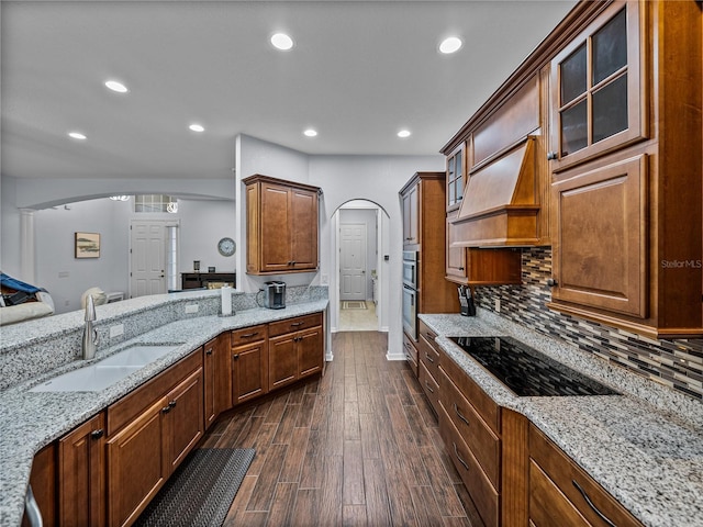 kitchen with arched walkways, custom range hood, a sink, black electric cooktop, and backsplash