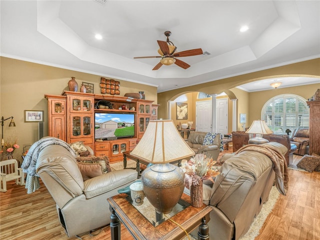 living room featuring arched walkways, a ceiling fan, light wood finished floors, ornamental molding, and a tray ceiling