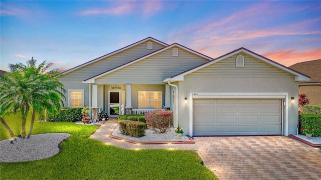 single story home with an attached garage, covered porch, decorative driveway, a lawn, and stucco siding