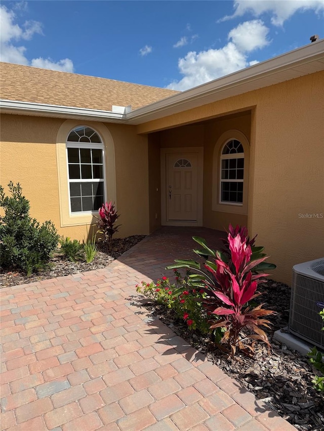 entrance to property with a shingled roof, cooling unit, and stucco siding