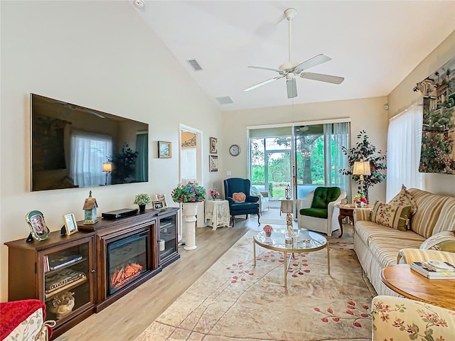 living room with light wood finished floors, visible vents, a glass covered fireplace, ceiling fan, and high vaulted ceiling