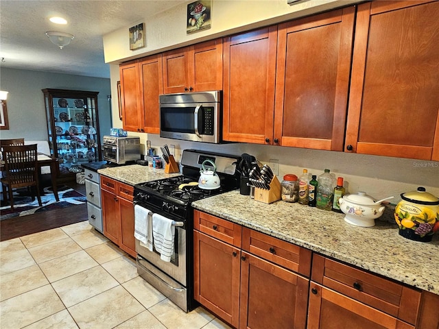 kitchen featuring brown cabinets, light tile patterned floors, appliances with stainless steel finishes, a textured ceiling, and light stone countertops