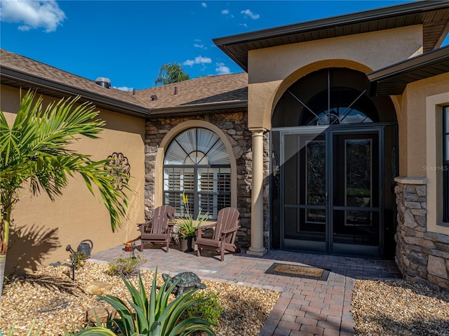 entrance to property featuring stone siding, roof with shingles, a patio, and stucco siding