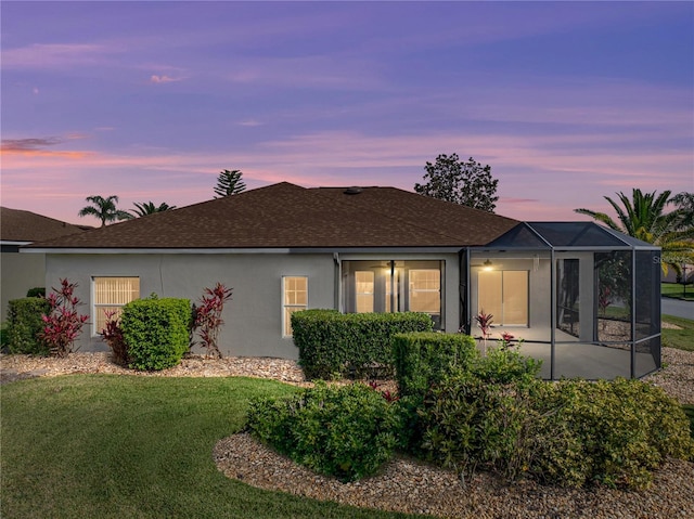 back of house at dusk featuring a lanai, a shingled roof, a lawn, and stucco siding