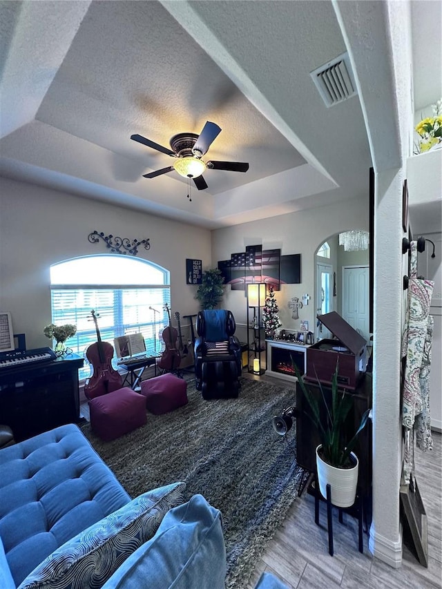 living room featuring a ceiling fan, visible vents, a tray ceiling, arched walkways, and a textured ceiling