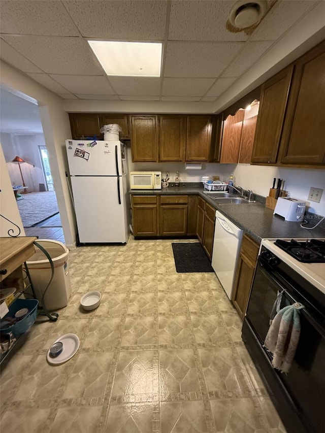 kitchen with white appliances, dark countertops, a paneled ceiling, light floors, and a sink