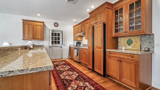 kitchen with stainless steel stove, under cabinet range hood, light wood-type flooring, brown cabinets, and glass insert cabinets