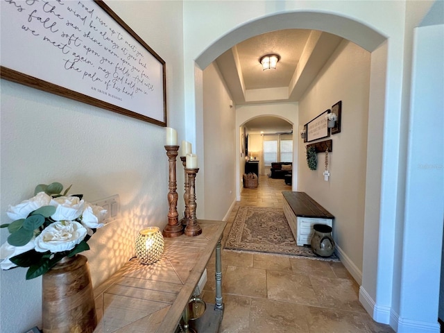 corridor featuring a raised ceiling, stone tile floors, baseboards, and arched walkways