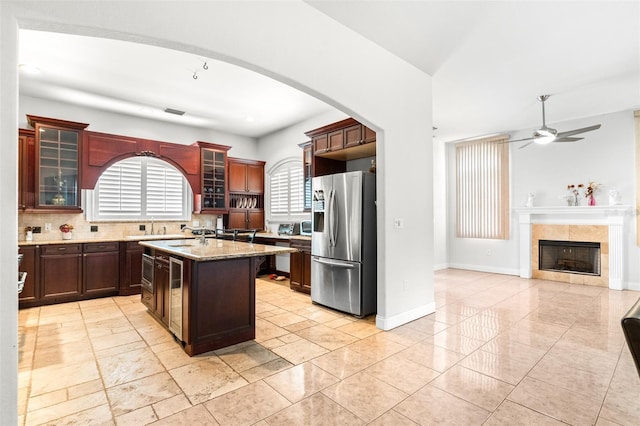 kitchen with light stone counters, a center island with sink, a ceiling fan, a tile fireplace, and stainless steel fridge