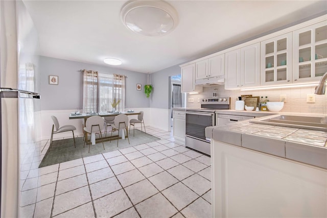 kitchen featuring a wainscoted wall, tile countertops, a sink, stainless steel range with electric stovetop, and under cabinet range hood