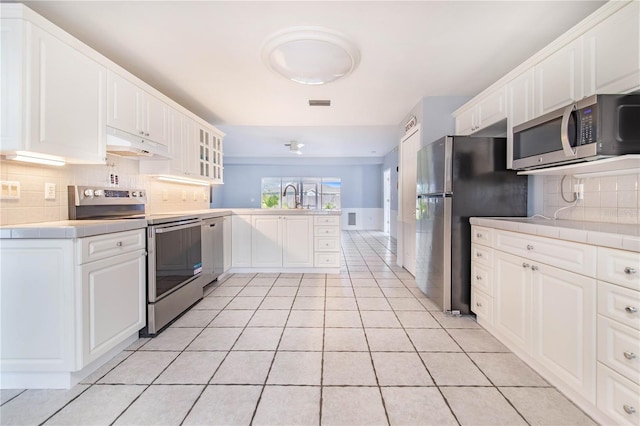 kitchen featuring tile countertops, light tile patterned flooring, under cabinet range hood, a peninsula, and appliances with stainless steel finishes