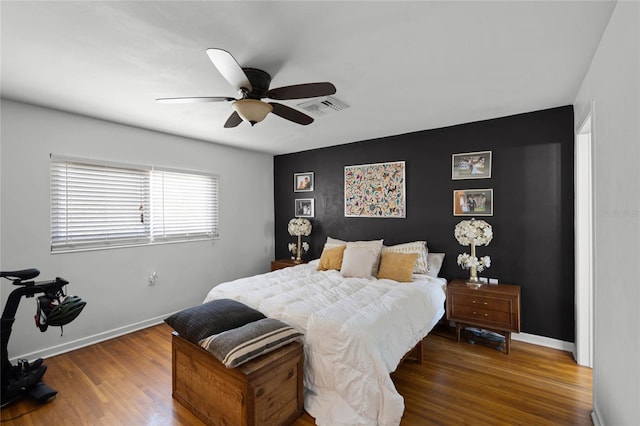 bedroom featuring visible vents, a ceiling fan, wood finished floors, baseboards, and an accent wall