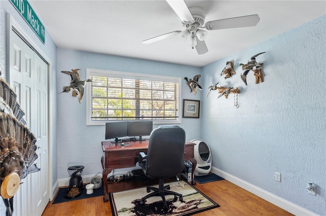 home office with a textured wall, wood finished floors, a ceiling fan, and baseboards