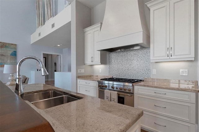 kitchen with backsplash, high end stove, premium range hood, white cabinets, and a sink