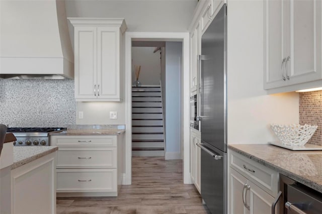 kitchen featuring light wood-type flooring, custom range hood, tasteful backsplash, appliances with stainless steel finishes, and white cabinets