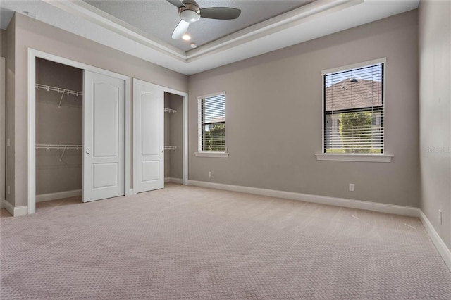 unfurnished bedroom with light carpet, two closets, a ceiling fan, a tray ceiling, and baseboards