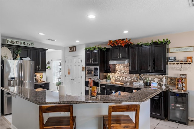 kitchen with under cabinet range hood, stainless steel appliances, wine cooler, decorative backsplash, and dark cabinets