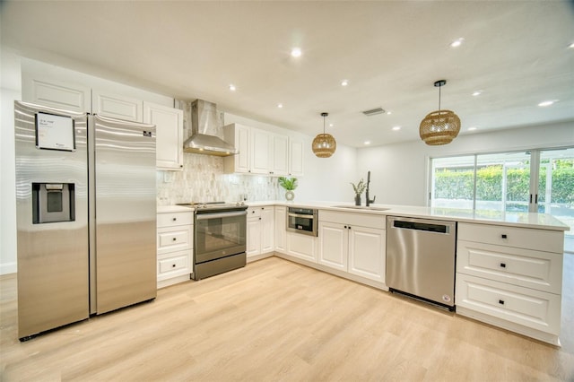kitchen with wall chimney range hood, a sink, light wood-type flooring, a peninsula, and appliances with stainless steel finishes
