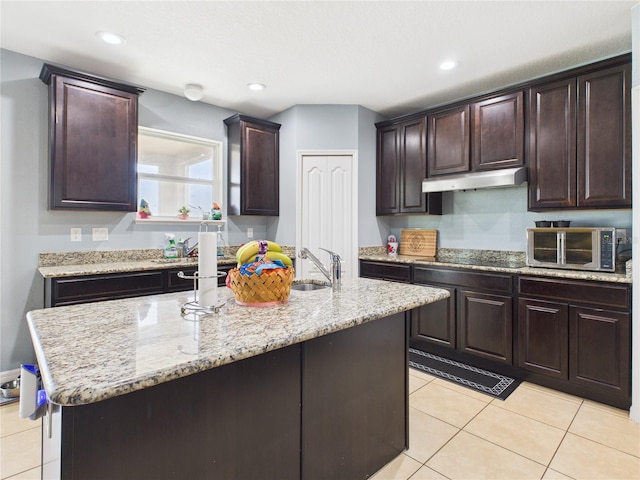 kitchen with a sink, under cabinet range hood, light tile patterned floors, a center island with sink, and dark brown cabinets