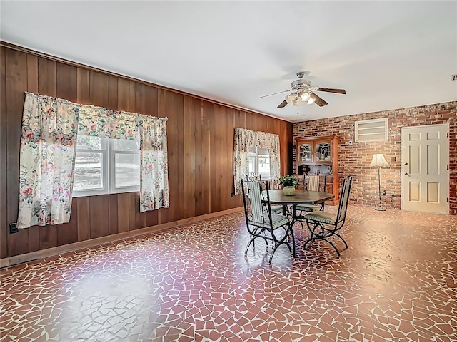 dining space with wooden walls, a ceiling fan, baseboards, and brick wall