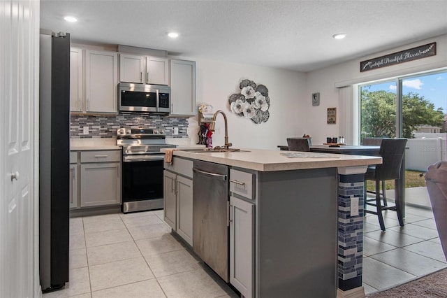 kitchen with appliances with stainless steel finishes, a sink, backsplash, gray cabinetry, and a center island with sink