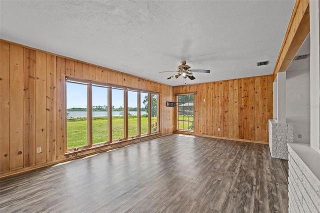 unfurnished living room featuring ceiling fan, visible vents, a water view, wood walls, and wood finished floors