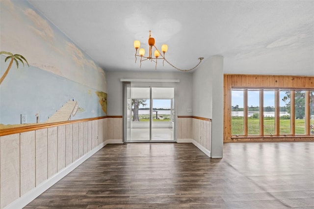 unfurnished dining area featuring a wainscoted wall, a notable chandelier, wood walls, dark wood-style flooring, and baseboards