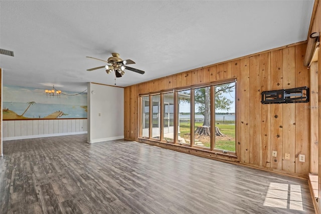 unfurnished living room with baseboards, visible vents, ceiling fan with notable chandelier, wooden walls, and wood finished floors
