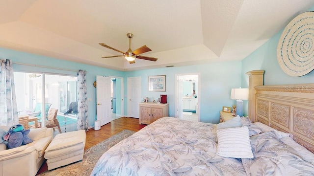 bedroom featuring ceiling fan, ensuite bathroom, access to outside, wood-type flooring, and a tray ceiling