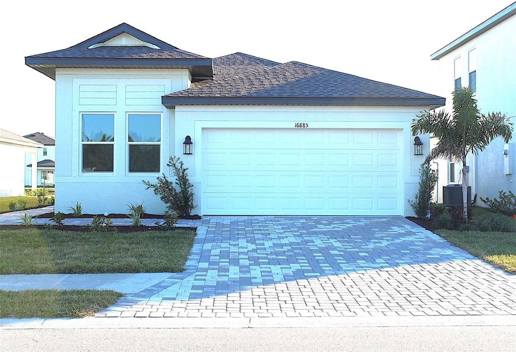 ranch-style house with a shingled roof, decorative driveway, an attached garage, and stucco siding