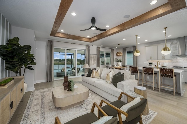 living room featuring baseboards, a raised ceiling, ceiling fan, light wood-style flooring, and recessed lighting